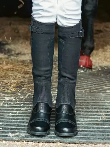 Front view of child standing in stable doorway wearing a pair of kids All Purpose neoprene half chaps in black by Just Chaps